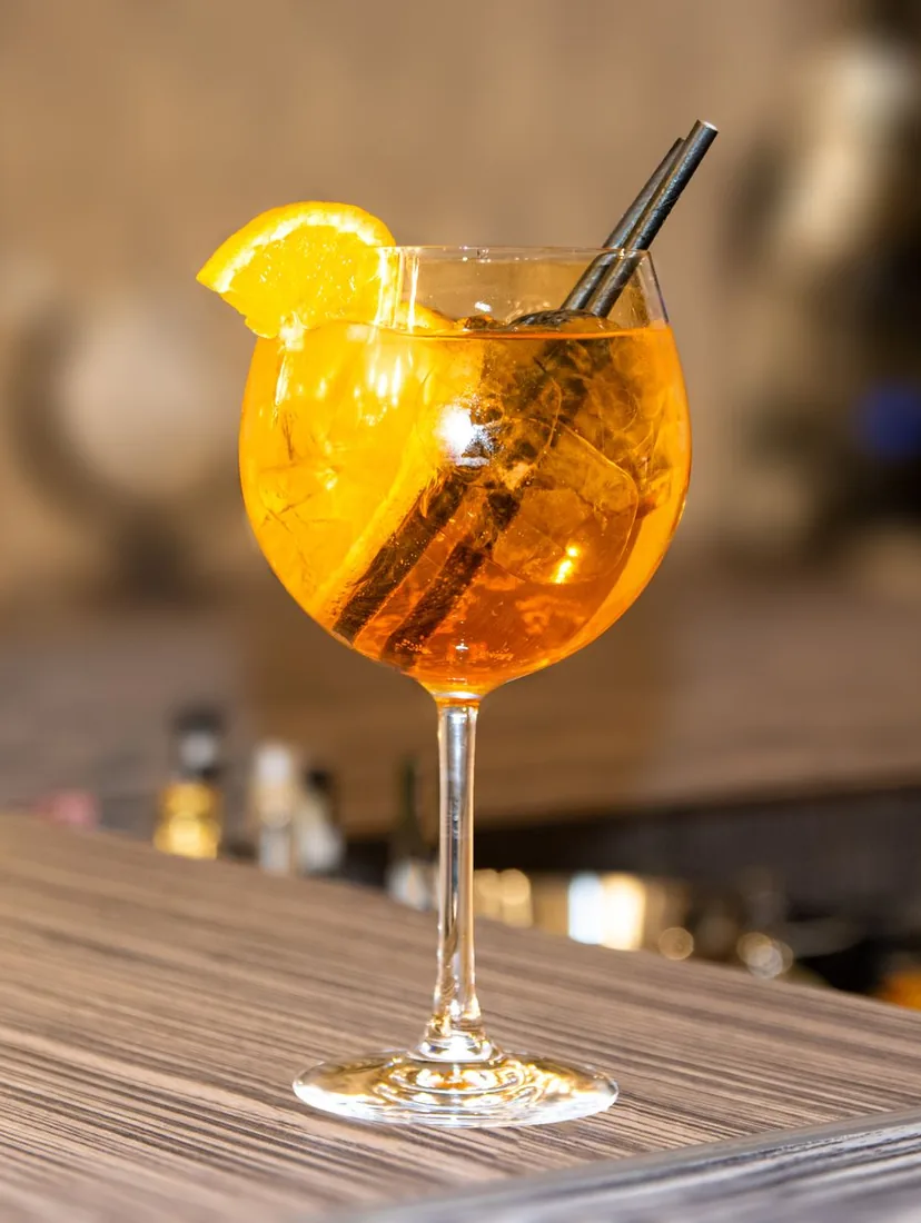A stemmed glass of orange spritz with an orange slice and a straw on a wooden bar counter