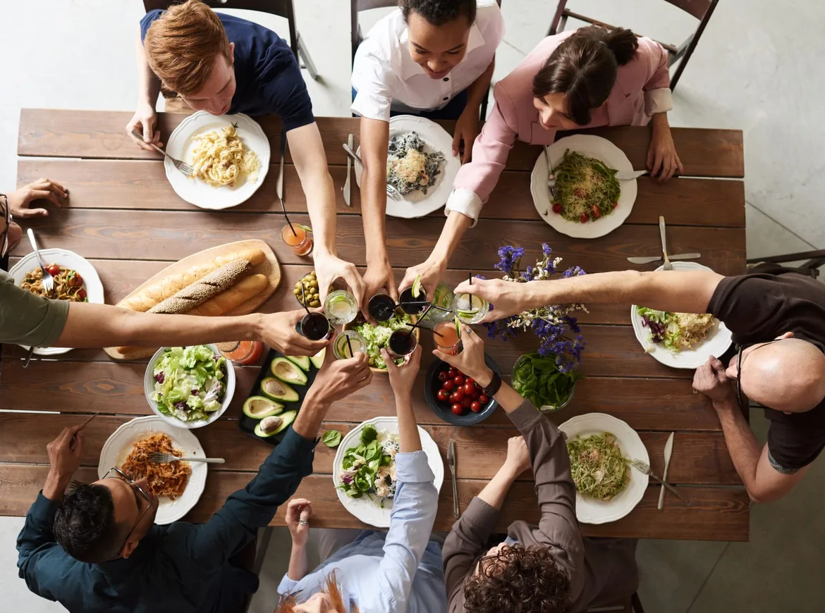 Seen from above, friends at a wooden table clink glasses over plates of pasta and salad